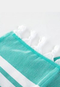 Turkusowo-biała plażowa ręcznik w paski z wzorem jodełki, z białymi frędzlami wzdłuż jednego brzegu. Miękka, tkana tekstura.