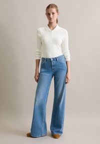 Weißes langärmeliges, geripptes Polo mit einem Halb-Reißverschluss-Kragen, kombiniert mit hellblauen, weit geschnittenen Jeans mit Fronttaschen und sichtbarer Naht.