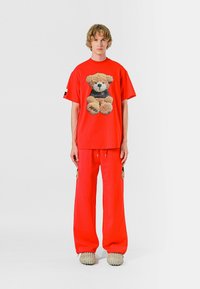 Camiseta de algodón roja con un gráfico de oso de peluche, combinada con pantalones de chándal rojos a juego. Tejido texturizado y diseño de corte holgado. Calzado casual.