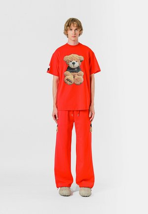 Camiseta de algodón roja con un gráfico de oso de peluche, combinada con pantalones de chándal rojos a juego. Tejido texturizado y diseño de corte holgado. Calzado casual.