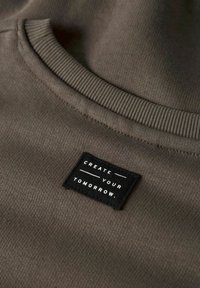 Närbild av en brun crewneck-tröja som visar en svart etikett med vit text "SKAPA DIN FRAMTID." sydd nära halslinningen.