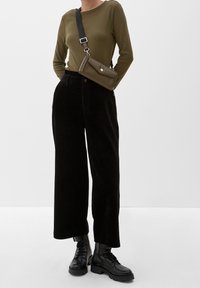 Camicia a maniche lunghe color olive abbinata a pantaloni larghi in corduroy neri e stivaletti neri con lacci, completata da una piccola borsa a tracolla verde.