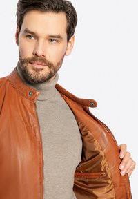 Giacca in pelle marrone con colletto a pressione e chiusura con zip, caratterizzata da una texture liscia e una tasca interna, abbinata a un dolcevita beige.