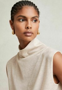 Beige ärmlös topp med en hög, vikbar krage gjord av mjukt tyg, som har en asymmetrisk söm och kompletteras av guldörhängen i form av hoop.