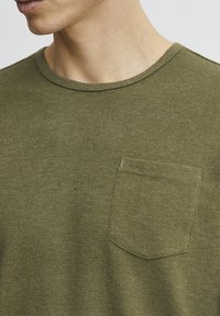 Olive green short-sleeve shirt, soft fabric with a round neckline and a chest pocket. Features a subtle texture and stitched accents.