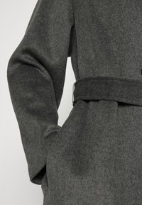 Grå uldblandingsfrakke med bælte i taljen, der har en blød tekstur og strukturerede ærmer. Den har knaplukning og et minimalistisk design.