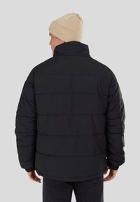 Doudoune noire avec des coutures horizontales, col haut, poignets côtelés et coupe décontractée. Portée avec un bonnet en tricot beige.