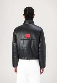 Czarna kurtka puchowa o błyszczącej fakturze, krótki fason, z przeszywanymi panelami. Z tyłu znajduje się czerwony prostokątny naszywka z logo.
