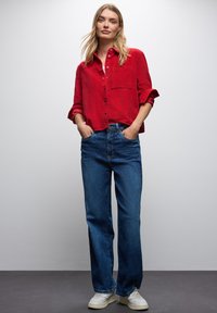 Rotes Hemd mit Knopfleiste aus strukturiertem Stoff und Tasche, kombiniert mit blauen, lässig geschnittenen Jeans und weißen Sneakers.