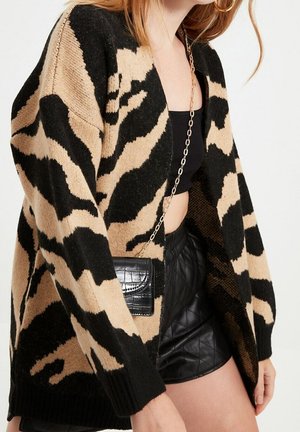 Frau trägt eine Strickjacke mit schwarzem und beigem Tiermuster, ein schwarzes Crop-Top, schwarze gesteppte Leder-Shorts und eine kleine schwarze Handtasche mit Kettenriemen.