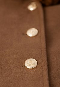 Brązowa filcowa kurtka ze złotymi guzikami, z detalem przedstawiającym herb. Zbliżenie guzika i tekstury materiału.