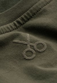 Olijfgroene katoenstof met een textuur geborduurd schaarontwerp. De halslijn heeft ribbeldetails voor extra textuur.