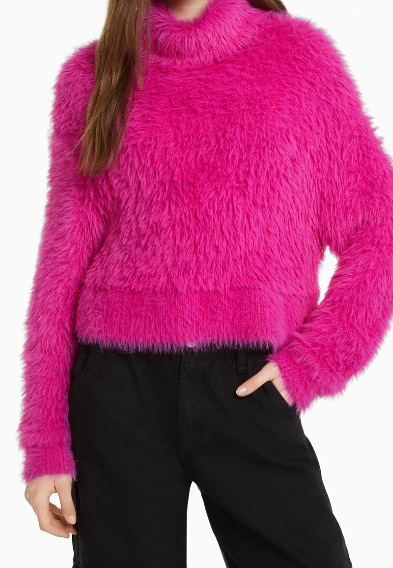 Flauschiger, hellrosa Pullover mit hohem Rollkragen, langen Ärmeln und cropped Design. Kombination mit schwarzen Hosen, die eine weiche Textur zeigen.