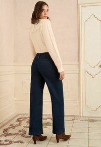 Puszysty kremowy sweter z koronkowym kołnierzykiem zestawiony z ciemnoniebieskimi, wysokim stanem spodniami o szerokich nogawkach oraz brązowymi czółenkami na szpic, na jasnym tle.