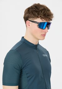 Cykeltrøje i mørk teal med teksturerede mesh-ærmer. Har en fuld lynlås foran og "ENDURANCE"-branding på brystet. Fremhæver blåtonede solbriller.