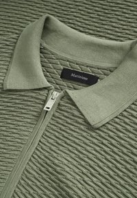 Gaiši zaļa teksturēta polo krekls ar rāvējslēdzi priekšpusē, ribaina apkakle un melna etiķete ar zīmola nosaukumu "Matinique".