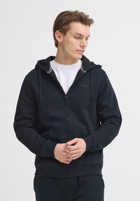 Sweat à capuche noir avec fermeture éclair, fabriqué en tissu doux. Comprend une capuche à cordon, des poches avant et un détail de logo subtil. Fini par des poignets et un ourlet côtelés.