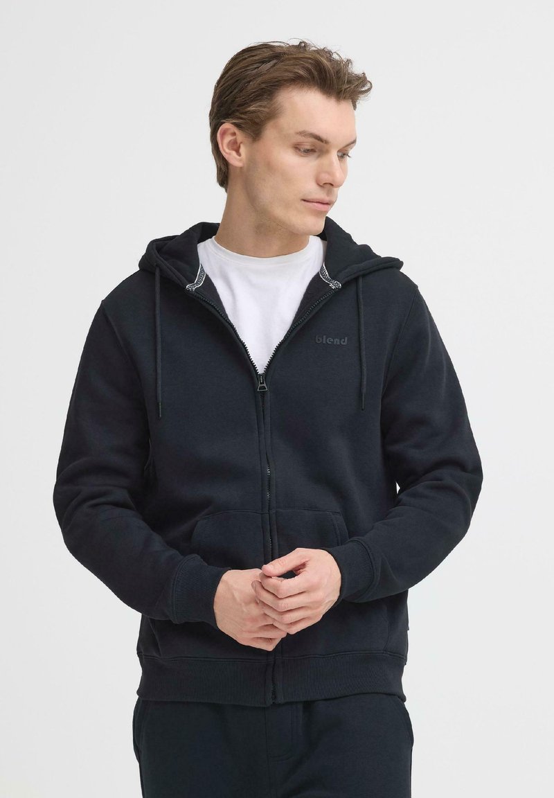 Sweat à capuche noir avec fermeture éclair, fabriqué en tissu doux. Comprend une capuche à cordon, des poches avant et un détail de logo subtil. Fini par des poignets et un ourlet côtelés.