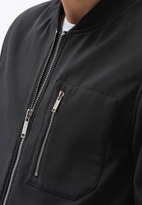 Black bomber jacket with a smooth texture, featuring a stand-up collar, two zippered pockets, and metallic zipper details.