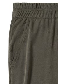 Gros plan sur un pantalon vert olive montrant une taille élastique et une poche latérale avec des coutures détaillées sur un tissu texturé.