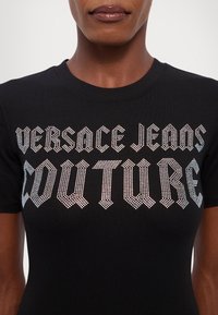 Čierne tričko s potlačou "VERSACE JEANS COUTURE" z kovových kamienkov. Krátke rukávy a klasický okrúhly výstrih. Hladká textúra látky.