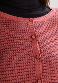 Texturerad, ribbade cardigan i rosafärg med runda, strukturerade knappar och rund halsringning, bärs över en svart polotröja.