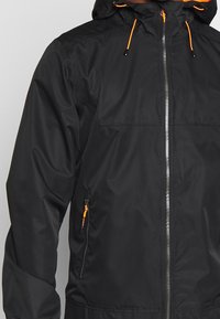 Sort jakke med lynlås foran og justerbar hætte. Har orange detaljer og en sidelomme med lynlås. Lavet af glat, letvægtsmateriale.