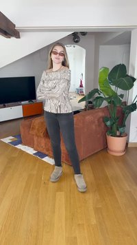 Top a maniche lunghe e spalle scoperte con un motivo astratto grigio e bianco, abbinato a jeans neri e stivali grigi. Ambientazione interna con piante verdi.