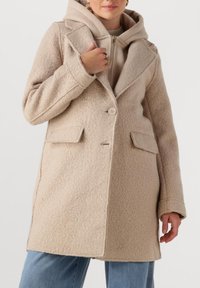 Beige wollenmix jas met een gestructureerde afwerking, grote revers, knoopsluiting met één knoop en twee voorzakken, gelaagd over een lichte hoodie.