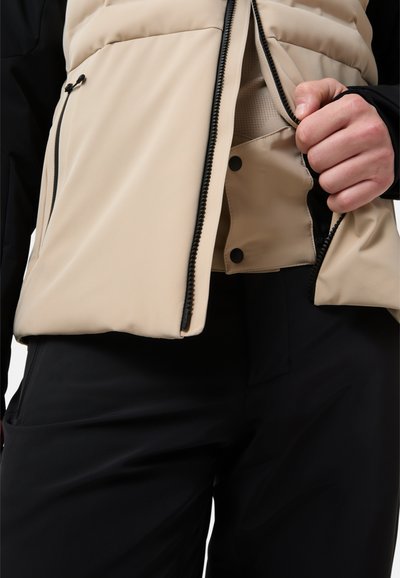 Beige gewatteerd vest, opengeknopt om een zwarte binnenkant te onthullen, met een gladde textuur en een contrasterende zwarte rits met drukknopsluiting. Zwarte broek zichtbaar.