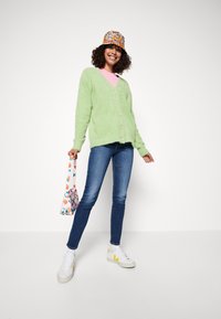 Πράσινο φουσκωτό κασκόλ, ροζ μπλούζα, μπλε τζιν και τσάντα με σχέδια. Τα αθλητικά παπούτσια είναι λευκά με κίτρινες λεπτομέρειες. Το μοντέλο ποζάρει χαμογελώντας.