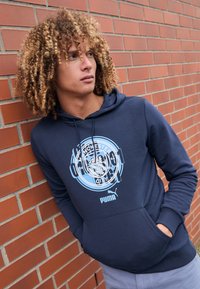 Hoodie blu navy con tasca a canguro, caratterizzato da una stampa del logo del Manchester City in azzurro chiaro e cordini regolabili. Composizione in misto cotone, texture liscia.