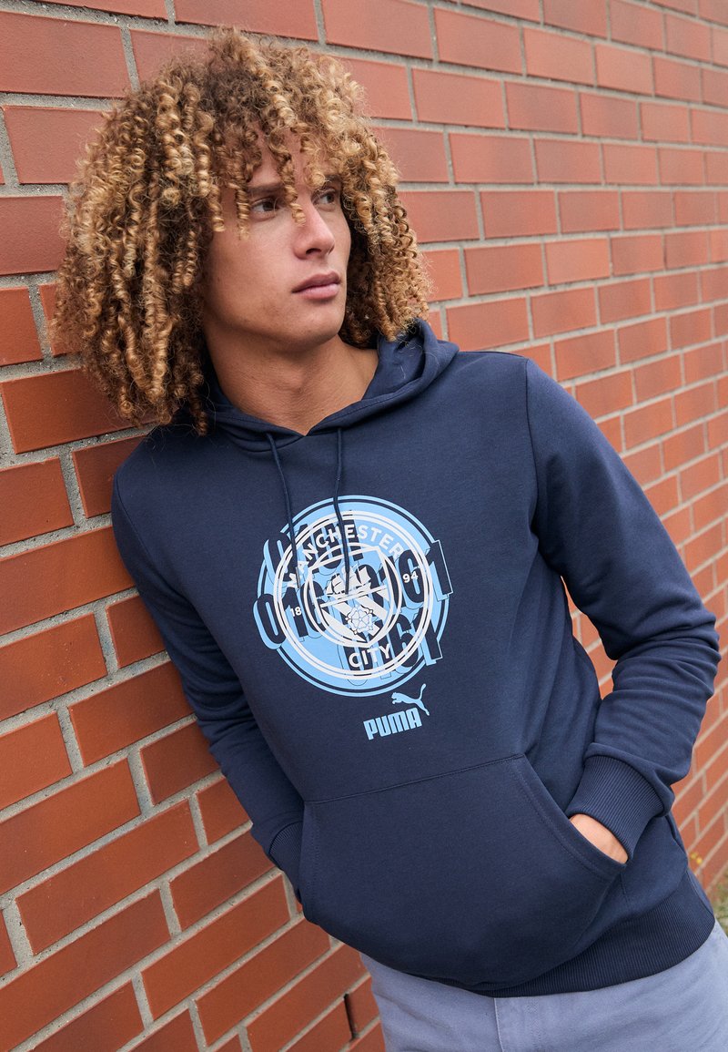 Hoodie blu navy con tasca a canguro, caratterizzato da una stampa del logo del Manchester City in azzurro chiaro e cordini regolabili. Composizione in misto cotone, texture liscia.