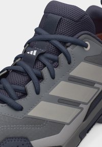 Dunkelblauer und grauer Sportschuh mit Netzzunge, überkreuzten Schnürsenkeln und drei hellgrauen Streifen an der Seite.