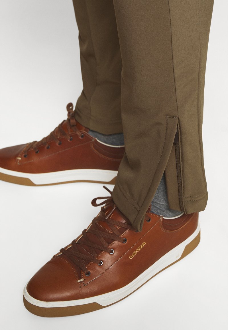 Baskets en cuir marron avec semelles blanches, associées à un pantalon kaki avec des zips sur le côté de l'ourlet, portées avec des chaussettes grises.