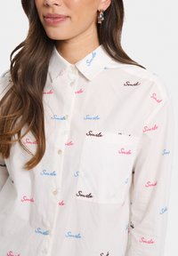 Camicia bianca con bottoni, caratterizzata da ricami colorati della scritta "Smile" in blu, rosa e marrone. Include una tasca sul petto e un design con colletto classico.