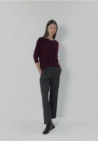 Burgundowy sweter z długim rękawem i ciemnoszare spodnie, charakteryzujące się luźnym krojem i gładką teksturą. Model stoi z rękami w kieszeniach na jednolitym tle.