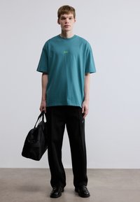Τυρκουάζ βαμβακερό t-shirt με στρογγυλή λαιμόκοψη και κοντά μανίκια· διαθέτει μικρό πράσινο λογότυπο. Συνδυάζεται με μαύρο άνετο παντελόνι και μαύρη τσάντα.