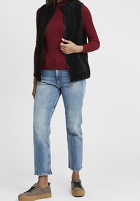Svart fleeceväst över en vinröd ribbad tröja, i kombination med ljusblå raka jeans och bruna rutiga sneakers.