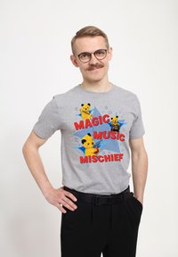 Šedé bavlněné tričko s krátkým rukávem, na kterém jsou tři grafiky karikatur medvědů a text "MAGICKÁ HUDBA ŠAŠKÁREN" tučnými červenými písmeny.