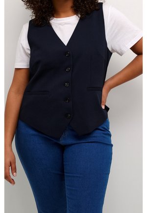 Frau trägt eine schwarze, geknöpfte Weste über einem weißen Kurzarmhemd und blauen Jeans, steht mit einer Hand in der Hüfte vor einem einfarbigen Hintergrund.