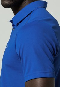 Homme portant un polo à manches courtes bleu vif avec un col, des coutures visibles et un petit logo blanc sur la poitrine.