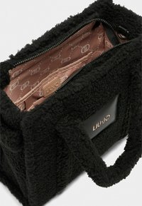 Sac textile noir et duveteux avec une texture douce, doté d'une fermeture éclair, d'un intérieur doublé rose et d'une étiquette logo noire sur le côté.