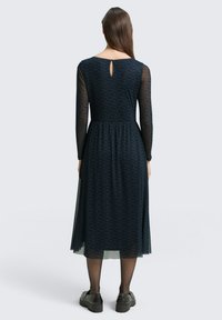 Navy jurk met lange doorzichtige mouwen, een keyhole-detail op de achterkant en een getailleerde taille. Een textuurpatroon bedekt de stof. Gemaakt met zwarte schoenen.