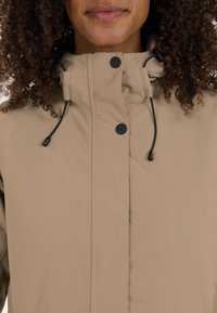 Beige jas met hoge kraag, voorzien van zwarte drukknoopsluitingen en trekkoordaccenten. Glad textuur met een minimalistisch ontwerp.