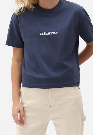 Osoba ubrana w granatowy krótki t-shirt z napisem "Dickies" oraz beżowe spodnie z wysokim stanem, stojąca z jedną ręką w kieszeni.