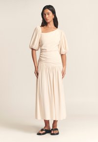 Beige midi-jurk met pofmouwen, vierkante halslijn en gathered taille. Vloeiende stofstructuur, gecombineerd met zwarte platte sandalen.