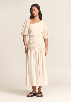 Beige Midi-Kleid mit Puffärmel, quadratischem Ausschnitt und gerafftem Bund. Fließende Stofftextur, kombiniert mit schwarzen flachen Sandalen.