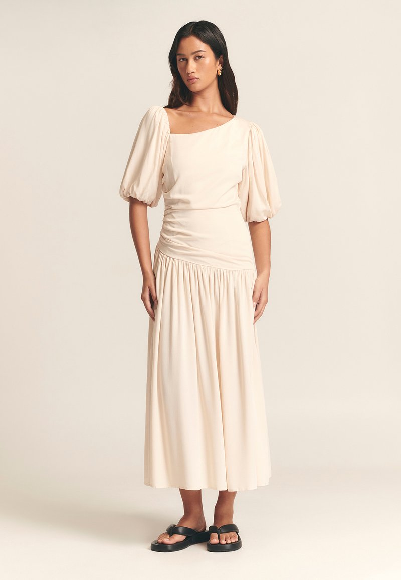 Beige midi-jurk met pofmouwen, vierkante halslijn en gathered taille. Vloeiende stofstructuur, gecombineerd met zwarte platte sandalen.