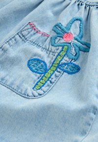 Tissu en denim avec une poche cousue ornée d'une fleur brodée colorée aux pétales bleus, au centre rose, à la tige verte et aux feuilles bleues.